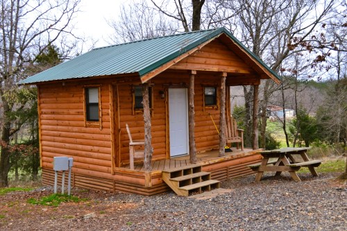 A small wooden cabin with a green metal roof, surrounded by trees and a gravel path, featuring a porch and picnic table.