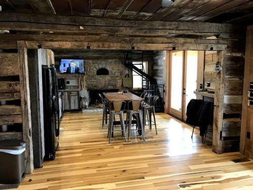 Cozy rustic kitchen with wooden beams, a dining table, and a TV, featuring warm wood floors and a stone wall.