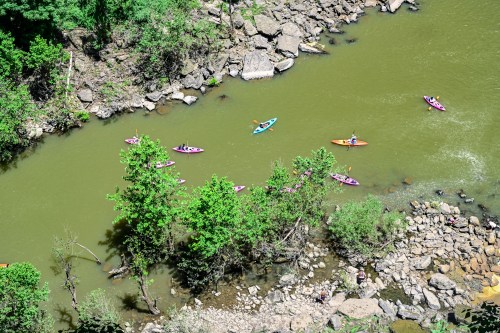 Aerial view of kayakers on a green river surrounded by rocky banks and lush trees.