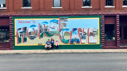 A family poses in front of a colorful mural that reads "Welcome to Horse Cave."