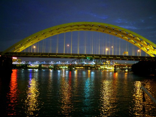 A brightly lit bridge arches over a river at night, reflecting colorful lights on the water's surface.