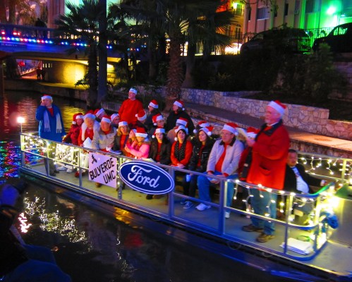 A festive group in Santa hats on a decorated boat, celebrating with lights along a canal.