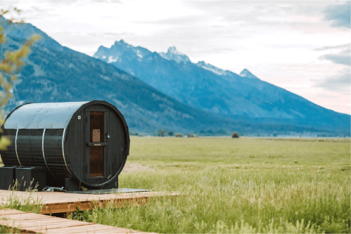A black barrel-shaped cabin on a wooden deck, set against a backdrop of mountains and a grassy field.