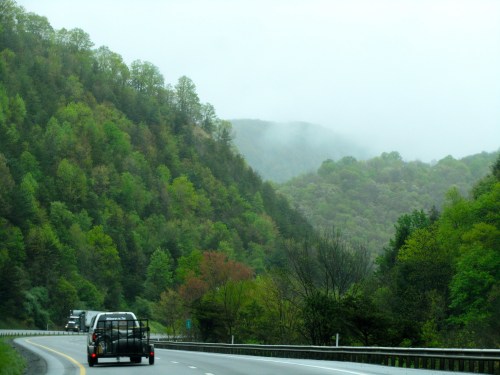 A truck towing a trailer drives along a winding road surrounded by lush green hills and misty mountains.