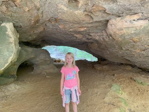 A young girl stands inside a cave, smiling, with rocky formations and a sandy floor around her.