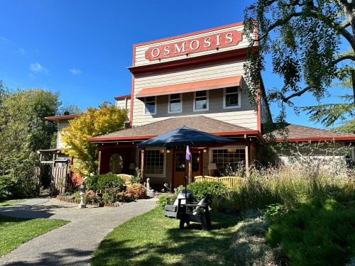A charming building with the sign "OSMOSIS," surrounded by greenery and a sunny blue sky.