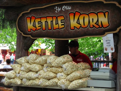 A vendor stand labeled "Ye Olde Kettle Korn" with bags of popcorn stacked in front and a cashier behind.