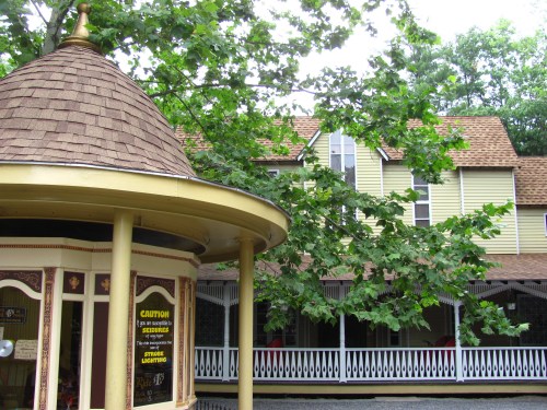 A gazebo with a shingled roof in front of a multi-story house surrounded by trees.