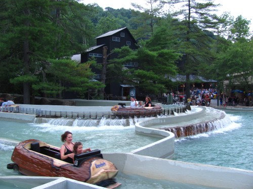 Two people enjoy a log flume ride in a water park, surrounded by trees and a busy building in the background.
