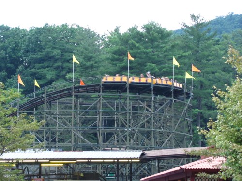 A wooden roller coaster with colorful flags, surrounded by trees, as riders enjoy a thrilling descent.