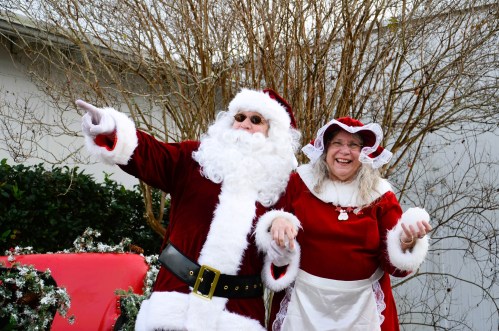 Santa Claus and Mrs. Claus joyfully pose together, with Santa pointing and both wearing festive holiday attire.