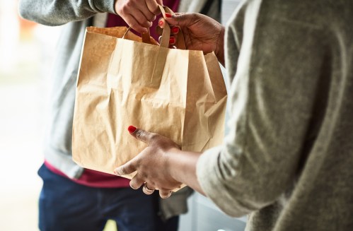 A person hands a brown paper bag to another person, both wearing casual clothing.
