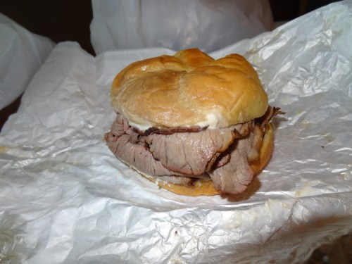 A close-up of a beef sandwich on a soft bun, resting on crumpled white paper.