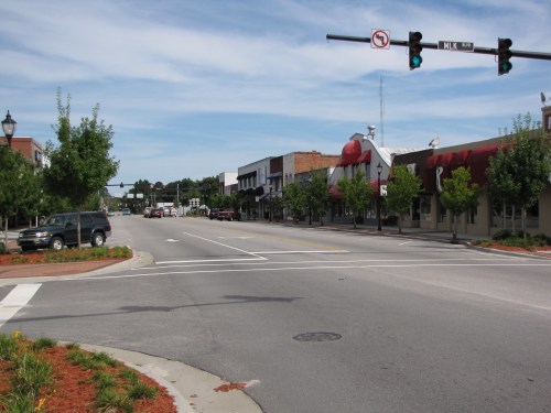 A quiet street scene with shops, traffic lights, and trees under a blue sky.