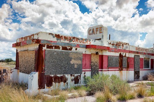 Abandoned café with boarded windows, peeling paint, and overgrown grass under a cloudy sky.