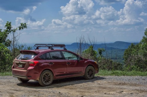 A dirt-covered Subura parked towards the view of the mountains on a gravel road.
