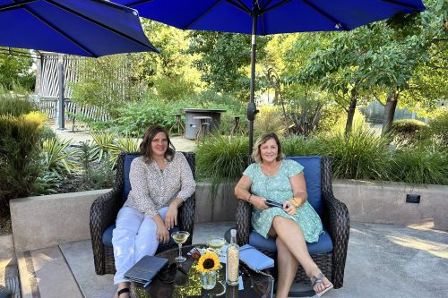 Two women sit together under a blue umbrella in a garden, enjoying drinks and smiling at the camera.