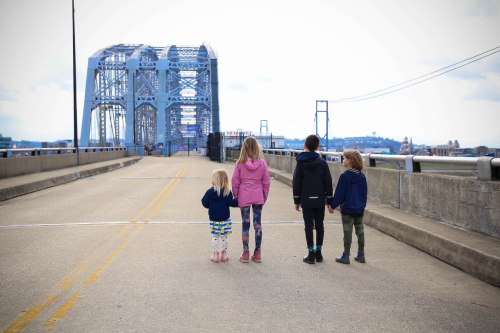 Four children stand on a bridge, facing away, with a large metal structure in the background and a cloudy sky above.