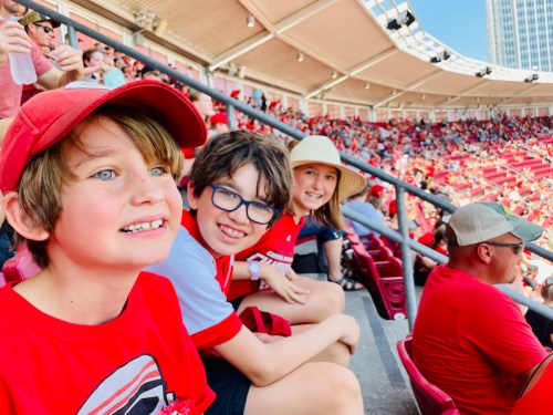 Three children in red shirts and hats smile while sitting in a stadium filled with fans in red.