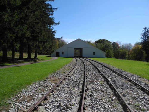 A white building with an open entrance, surrounded by green grass and trees, with railway tracks leading towards it.