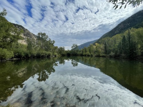 A serene landscape featuring a calm river reflecting clouds and trees, surrounded by mountains and greenery.