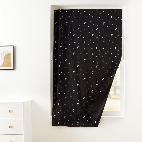 Black curtain with white crescent moon and star pattern, hanging in a window with a light-colored wall and furniture nearby.