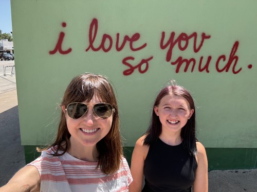 Two smiling women pose in front of a green wall with red graffiti that says, "I love you so much."