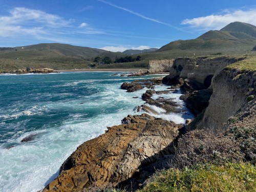 Coastal view with rocky cliffs, crashing waves, and rolling green hills under a blue sky with scattered clouds.