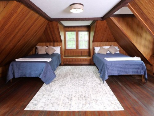 Cozy attic bedroom with two beds, wooden walls, and a patterned rug, featuring large windows for natural light.