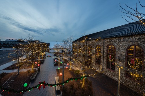 A festive outdoor scene with trees adorned in lights and decorations, alongside a stone building at dusk.
