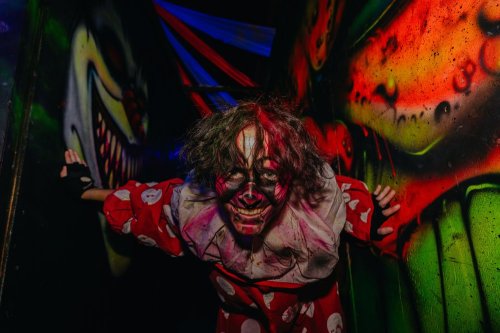 A creepy clown in a colorful costume leans against a wall, surrounded by vibrant, eerie graffiti.