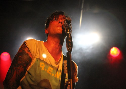 A singer with tattoos performs on stage, illuminated by colorful lights, holding a vintage microphone.