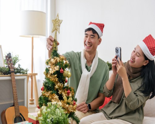 A joyful couple in cozy winter coats and scarves sharing a warm embrace and laughter under falling snow, surrounded by twinkling Christmas lights, a decorated evergreen tree