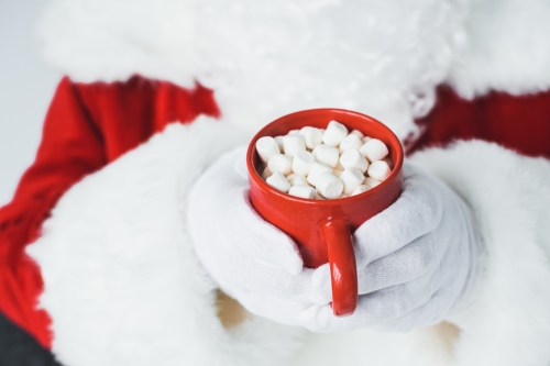 A close-up of Santa's hands holding a red mug filled with marshmallows. Santa is wearing a red suit with white fur trim.