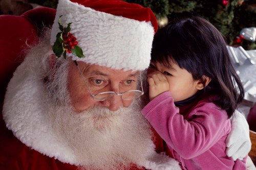 A young child whispers to Santa Claus, who has a white beard and glasses, in a festive setting.