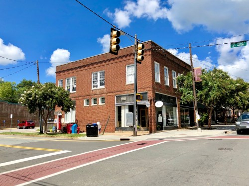 A brick building at a street corner with traffic lights, trees, and parked cars under a blue sky with clouds.