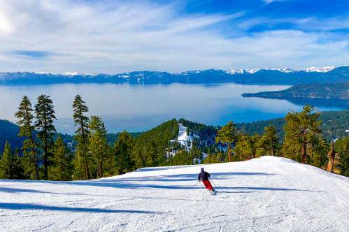 A skier descends a snowy slope with a stunning view of a lake and mountains in the background under a clear blue sky.