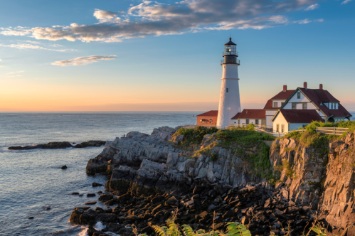 The sun rises over the Portland Head Light lighthouse in Cape Elizabeth, Maine. The lighthouse is situated on a rocky shore and the ocean is in view.