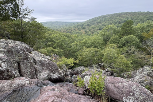 A rocky outcrop overlooks a lush green valley with rolling hills under a cloudy sky.