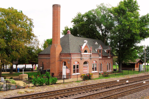 Historic brick train station with a tall chimney, surrounded by trees and a railway track in the foreground.