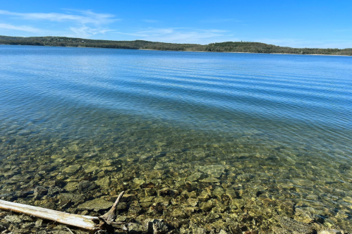 A calm lake with clear water revealing rocky bottom, surrounded by green hills under a blue sky.