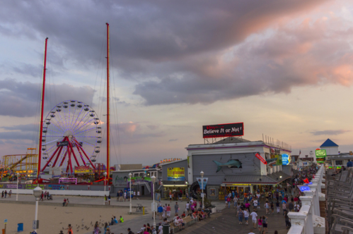 View of the Ocean City Boardwalk and beach at sunset in Ocean City, Maryland. People walk down the boardwalk. A Ferris wheel and roller-coaster are in the background.