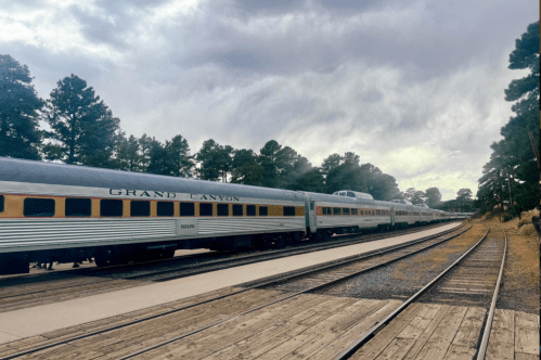 A vintage train labeled "Grand Canyon" parked on wooden tracks surrounded by pine trees under a cloudy sky.