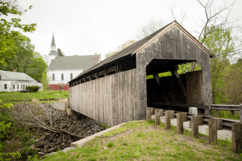Angled view of the side and front of the Burkeville Covered Bridge in Conway, MA. The wooden bridge is in the forefront and a white church is in the background.