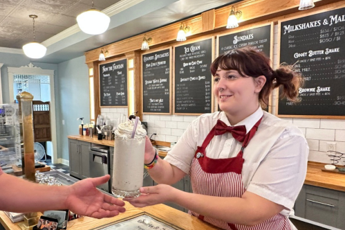 A smiling server in a bow tie hands a milkshake to a customer at a vintage ice cream shop.