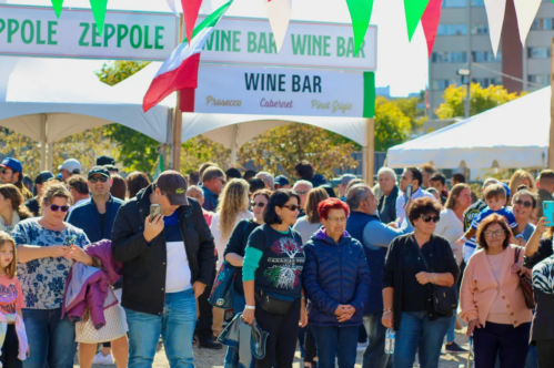 A crowd of people at an outdoor event, with tents labeled "Wine Bar" and an Italian flag in the background.