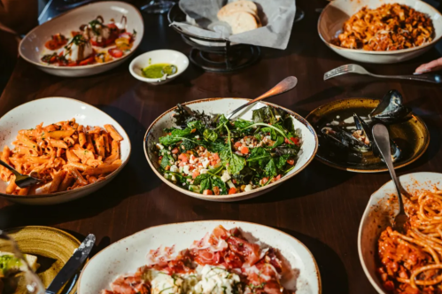 A table spread with various dishes, including pasta, salad, and seafood, surrounded by plates and utensils.