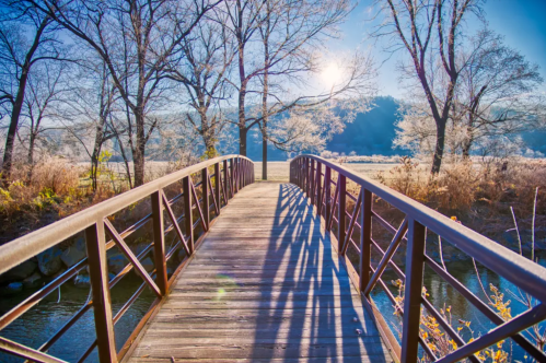A wooden bridge spans a serene landscape, surrounded by frosty trees and a bright sun shining in a clear blue sky.