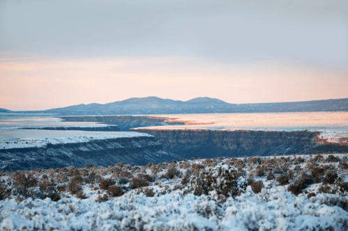 A snowy landscape with distant mountains and a canyon under a soft, pastel sky at dawn.