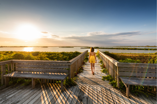 A girl in a yellow dress walks along a wooden boardwalk towards a sunset over a calm waterway.